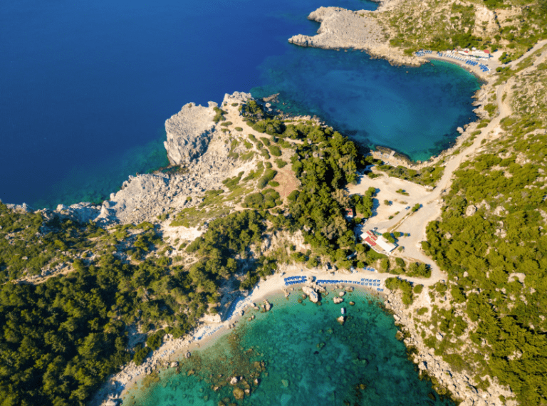 Aerial view of a rocky coastline with clear turquoise waters and scattered greenery. Small beaches boast lined umbrellas, embraced by the calm blue sea surrounding the coast. Roads and paths wind through the terrain, perfect for a My Dolce Vita Cruise or Rhodes Private Cruises experience.