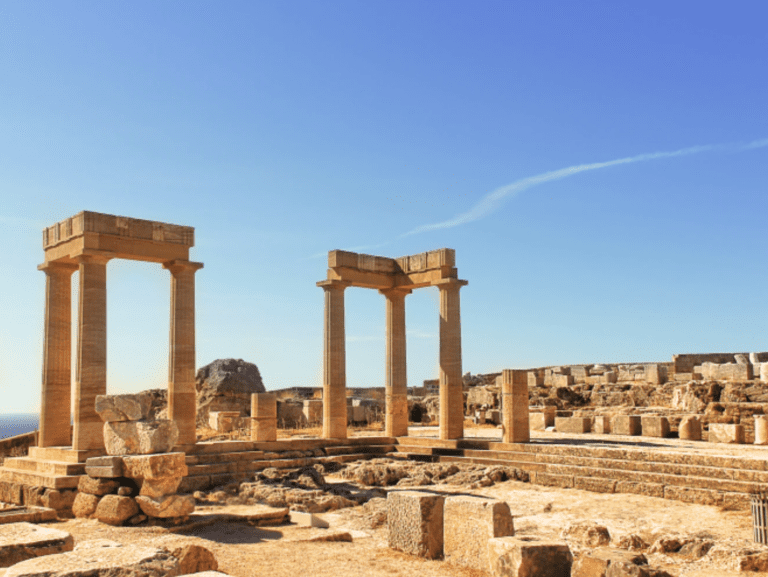 Amidst the ruins of an ancient Greek temple with standing columns under a clear blue sky, scattered stones hint at a rich history. As you gaze upon the barren landscape beyond, imagine embarking on a Rhodes Private Cruise to discover more of such timeless beauty.
