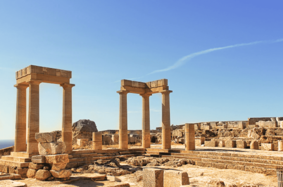 Amidst the ruins of an ancient Greek temple with standing columns under a clear blue sky, scattered stones hint at a rich history. As you gaze upon the barren landscape beyond, imagine embarking on a Rhodes Private Cruise to discover more of such timeless beauty.