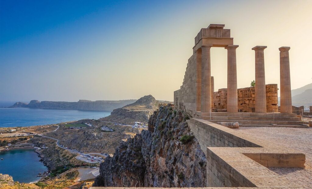 Ancient stone columns of the Lindos Acropolis stand on a rocky cliff, overlooking blue waters and a coastal landscape in Rhodes, Greece. Experience this enchanting view with Rhodes Private Cruises as the scene is illuminated by soft, golden light under a clear sky.