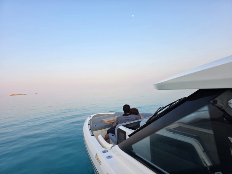A couple sits closely together at the bow of a white boat gliding over calm, clear blue water under an open sky with the moon visible, creating a peaceful and romantic atmosphere.