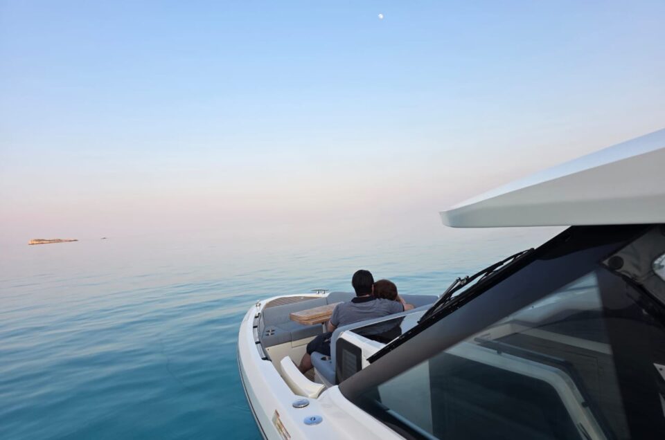 A couple sits closely together at the bow of a white boat gliding over calm, clear blue water under an open sky with the moon visible, creating a peaceful and romantic atmosphere.