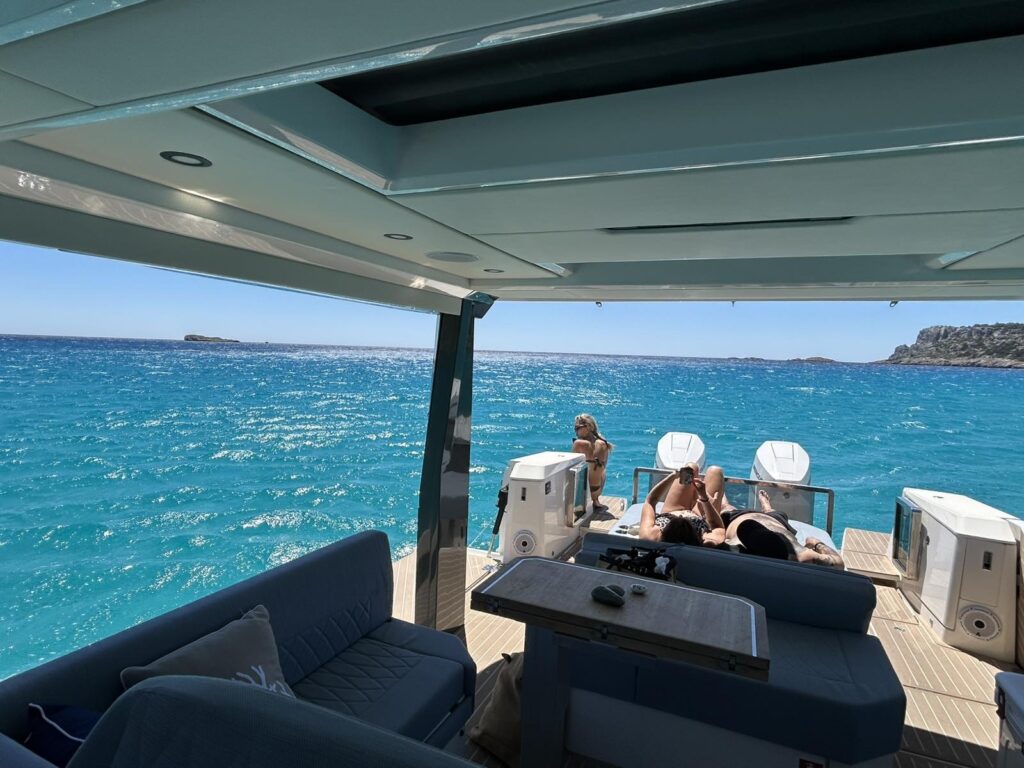 Two people relax on lounge chairs at the back of a boat, enjoying the sun and turquoise sea, with a third person sitting nearby. The boats seating area and table are visible in the foreground.