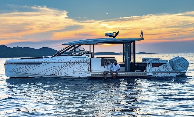 A modern white motor yacht floats on calm water at sunset, with two people sitting at the back, mountains in the distance, and a colorful sky reflecting on the sea.